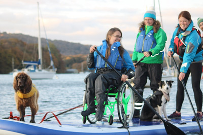A person in a wheelchair takes part in a paddleboard activity session. With them is two dogs and two Able 2 Adventure support staff members.