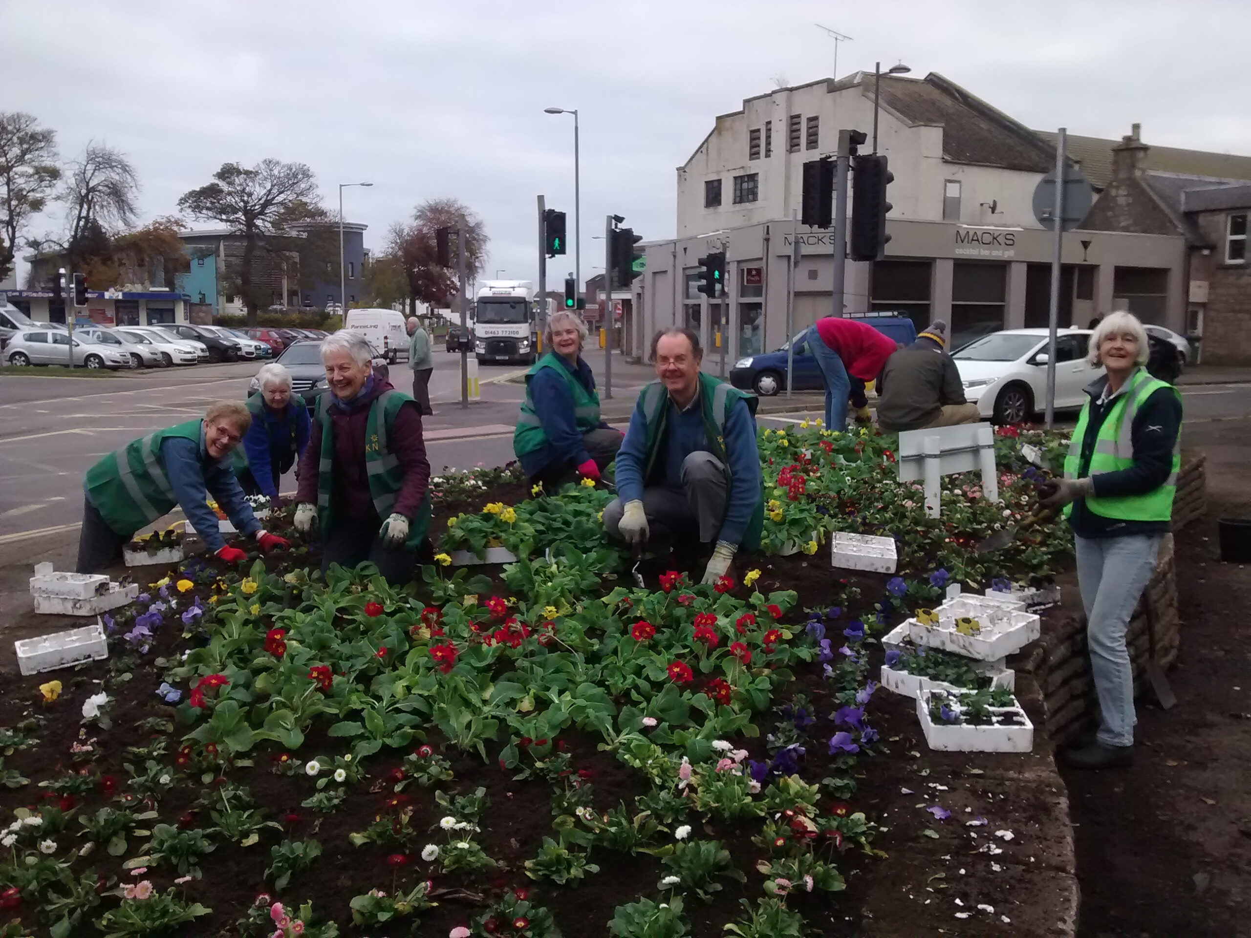 A group of people smiling at the photographer. They have been working on planting brambles in a bedding area outside in a pedestrian part or Nairn.