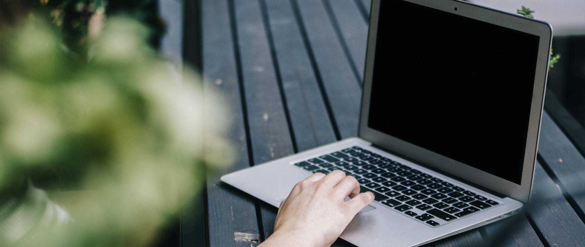 A laptop on a park bench. A person's hand is seen to be using it but the rest of them are out of the shot.