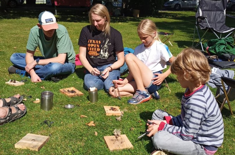 Children of various ages are sitting on a lawn with an adult, carrying out a handicraft activity.