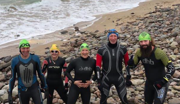 A group of outdoor swimmers, all in wetsuits and swimming caps, stand upon a stony beach, smiling at the camera.