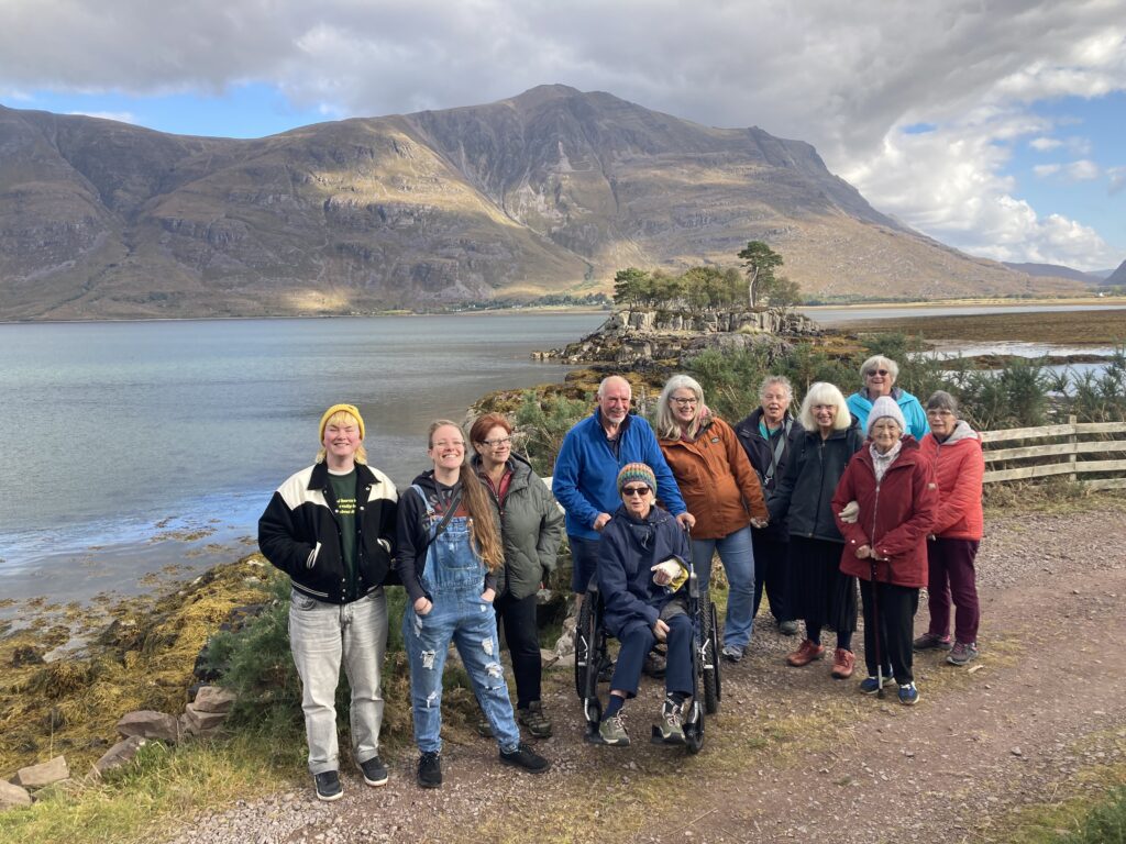 Group of smiling people standing on a path in front of a loch and mountain landscape. Group includes a wheelchair user.