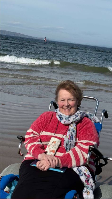 An older woman sitting in a specialized blue beach wheelchair, designed for navigating soft sand. The individual is smiling and looking towards the camera. They are on a beach with gentle waves and a cloudy sky. In the distance, a figure engaged in windsurfing can be seen on the water. The person in the wheelchair is wearing a red and white striped sweater, a dark skirt, and a patterned scarf.