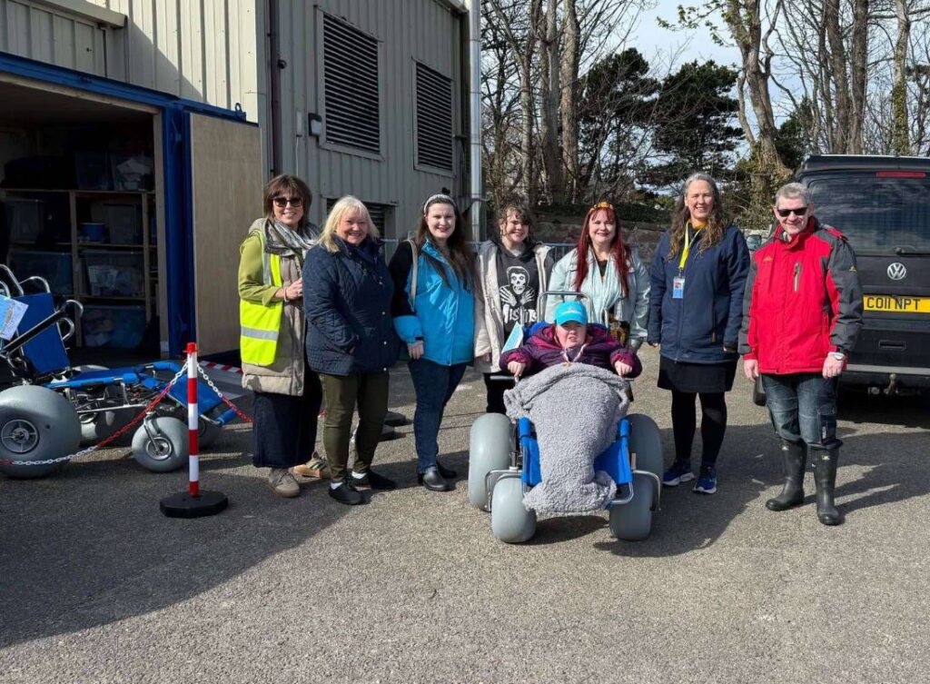 This photo shows seven people, representing the staff of the 'Nairn Beach Wheelchairs' group, gathered outside on a sunny day. In the center foreground, a person is seated in a specialized blue and grey beach wheelchair designed for sandy terrain. Six other adults are standing behind and to the sides of the wheelchair user, looking towards the camera. The background includes a building with a partially open storage unit, a parked vehicle, and some trees.