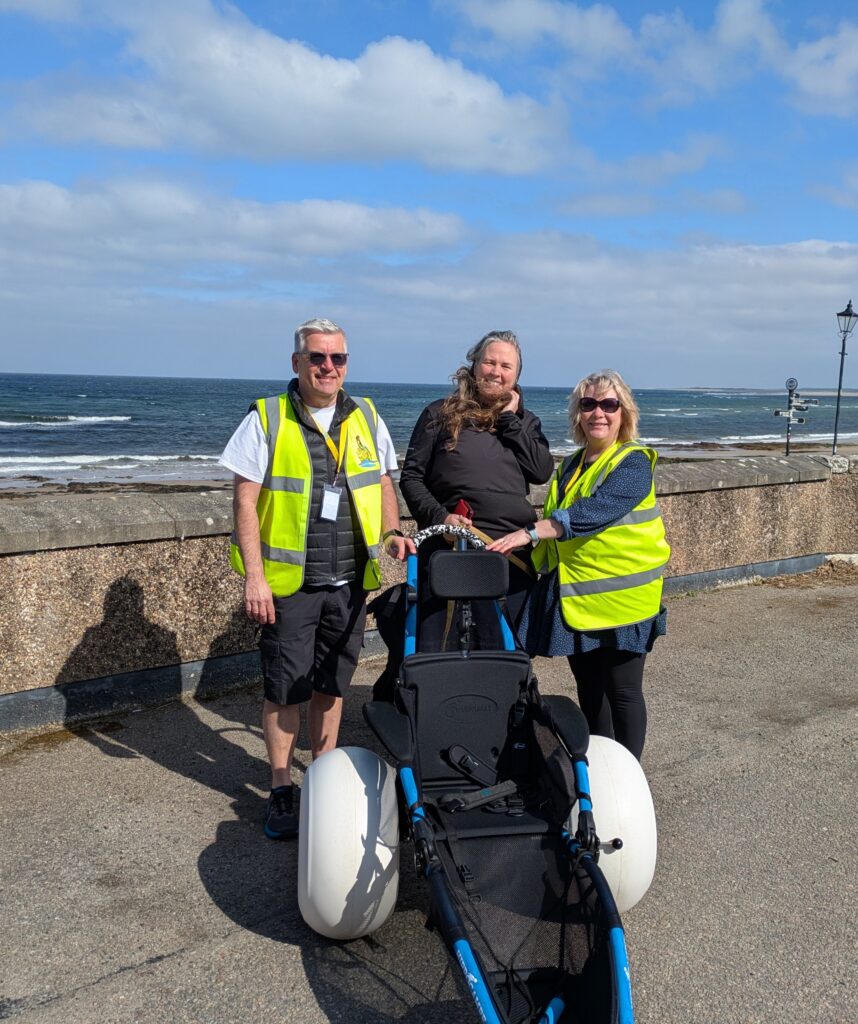 This image features three individuals positioned on a paved area, with a beach and ocean visible in the background. A specialised beach wheelchair, equipped with oversized, low-pressure tires for navigating sandy terrain, is prominently displayed in the foreground. Two of the individuals are wearing bright yellow high-visibility vests, suggesting they may be assisting with the wheelchair.