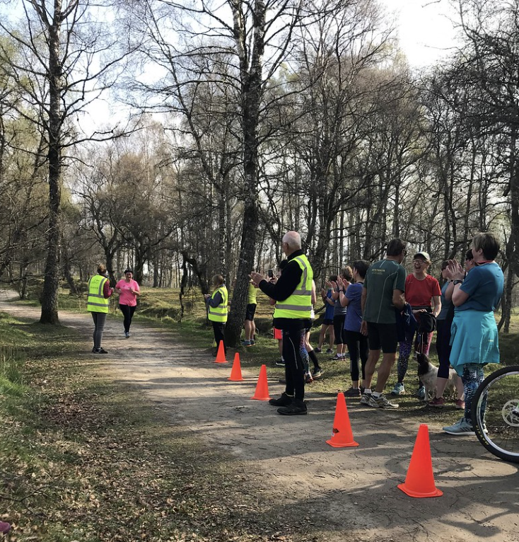 A person is running along a path in a rural location. There are stewards in high-vis vests applauding as the person runs past. There is also a small crowd of other runners chatting and applauding.