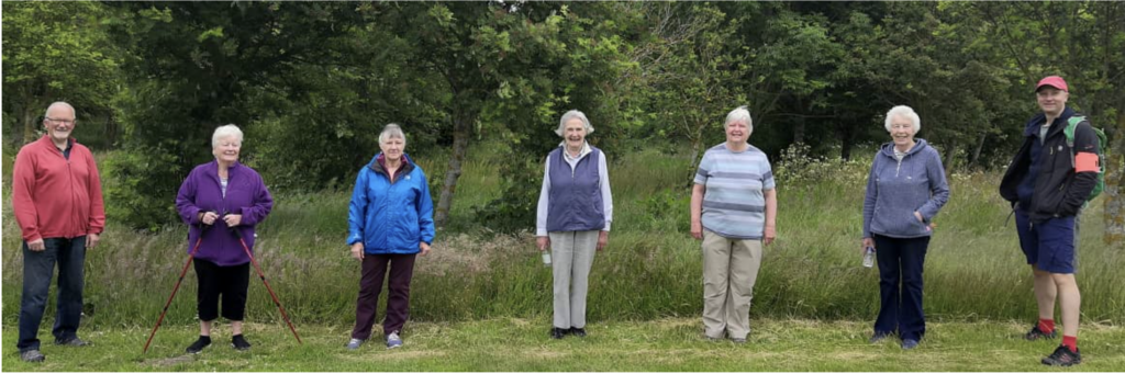 Group of older people lined up shoulder to shoulder smiling at the camera. They are stood in front of a woodland area on a lawn.