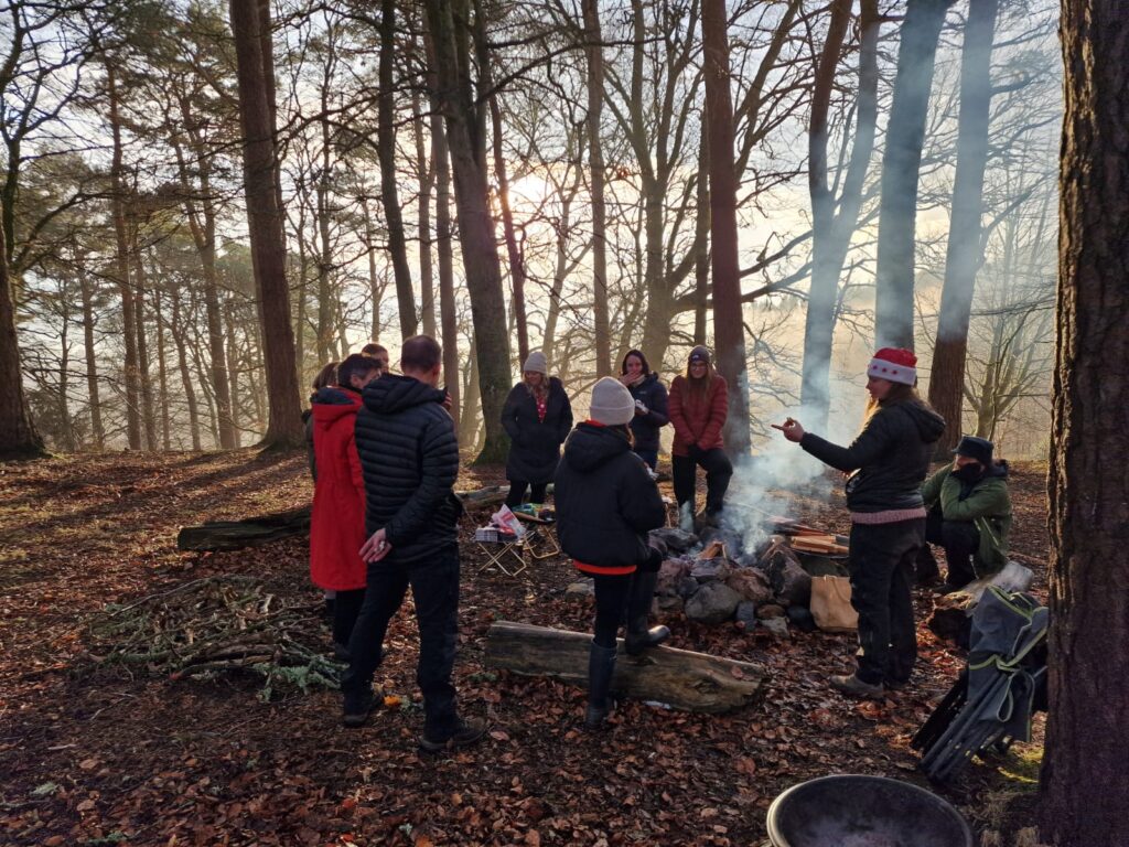 A group of people stand around a fire in the woods. Smoke is coming from the fire and people are chatting to each other