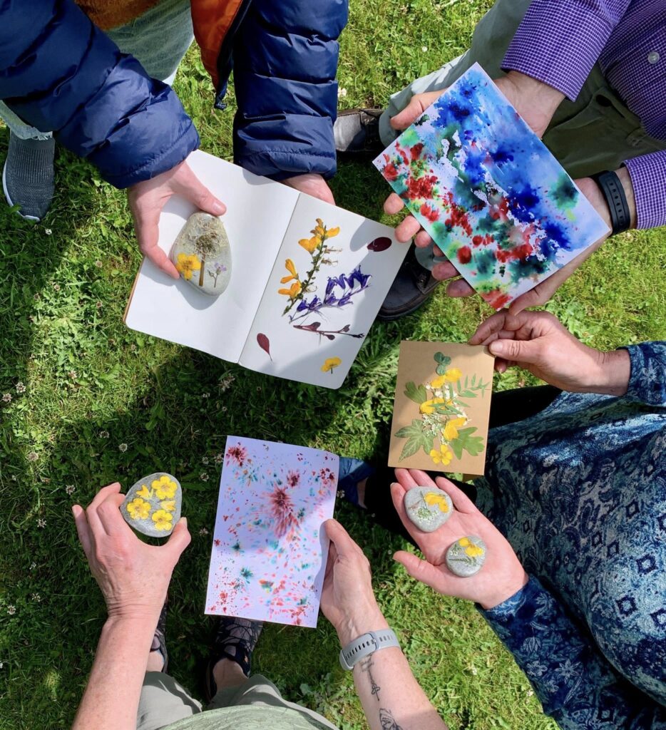 four people hands holding bright art work with flowers and abstract designs. Some people hold stones with small detailed art work on them