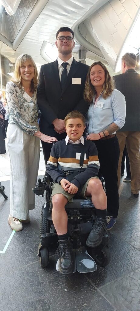 Three people stand smiling and one wheelchair user is smiling too.